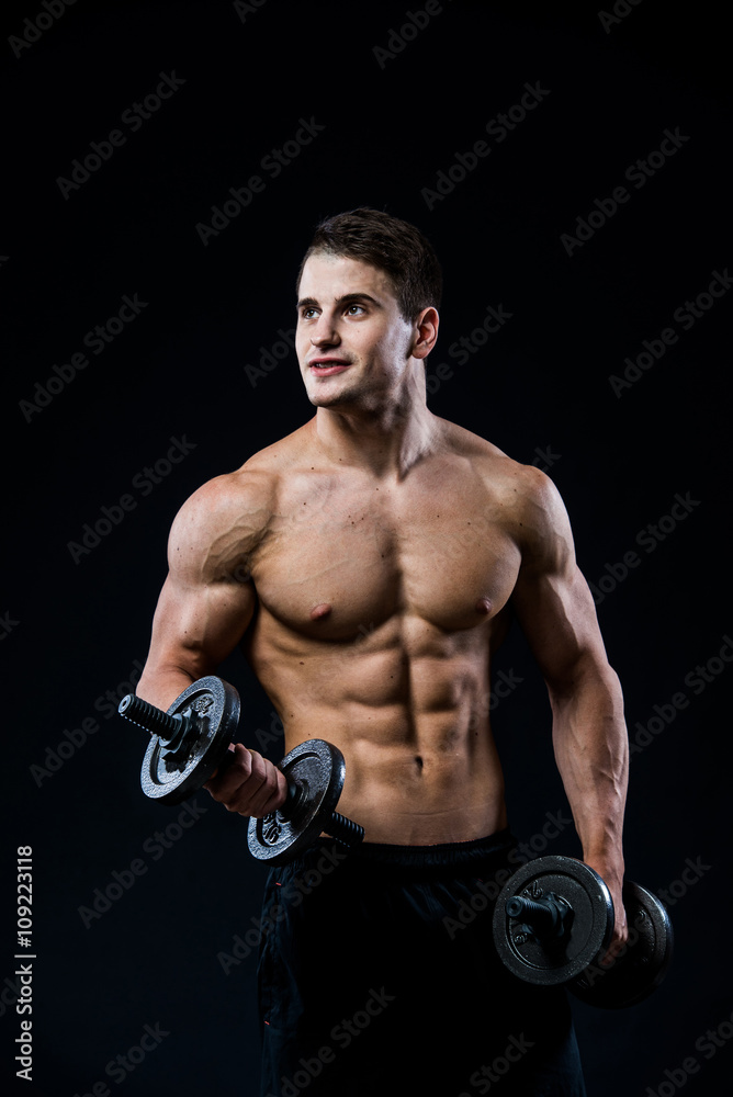 Young and fit male model posing his muscles pumping up with dumbbells in a gym looking to the left isolated on black background.