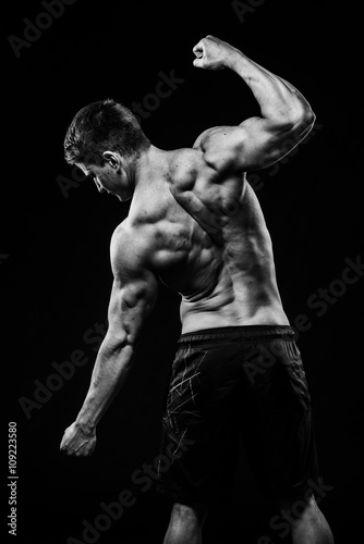 Young and fit bodybuilder athlete demonstrates biceps back view isolated on black background
