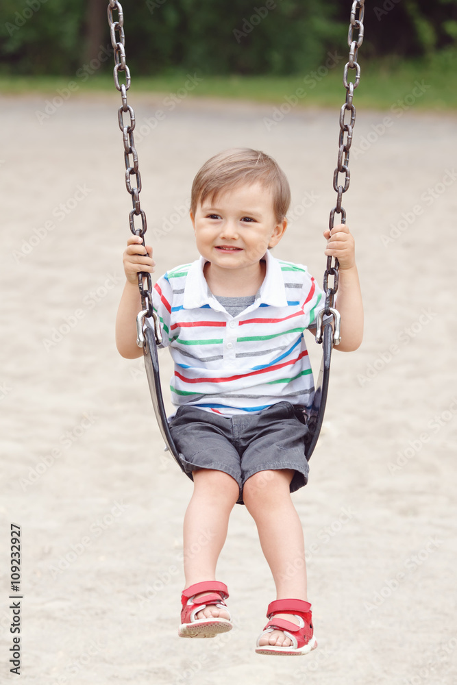 Portrait of happy smiling little boy toddler in tshirt and jeans shorts on swing on backyard playground outside on summer day, happy childhood lifestyle concept
