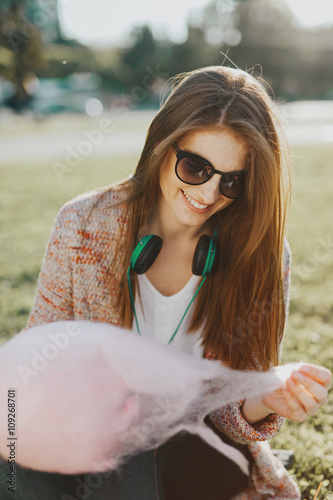 cute, beautiful caucasian white young lady looking enjoy relax in a green park and eats cotton candy while listens to the music with fashion style headphones