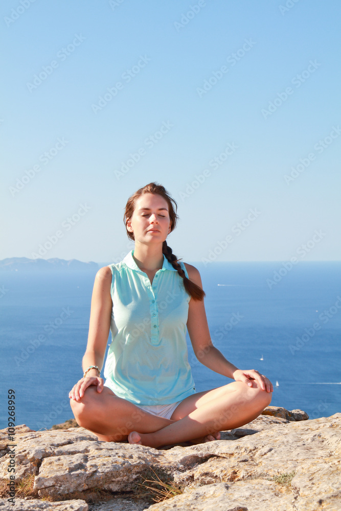 Beautiful young woman meditating outside on the top of the mountain with the blue sea behind her. Beautiful young woman meditating outside on the top of the mountain with the blue sea behind her.