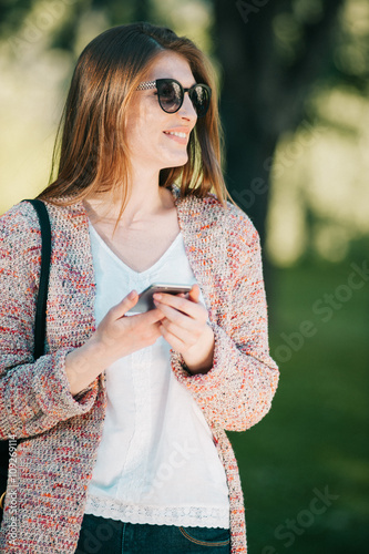 Cute caucasian young woman talking emotional and chat on the phone in the street near green park on spring sunset