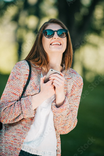 Cute caucasian young woman talking emotional and chat on the phone in the street near green park on spring sunset