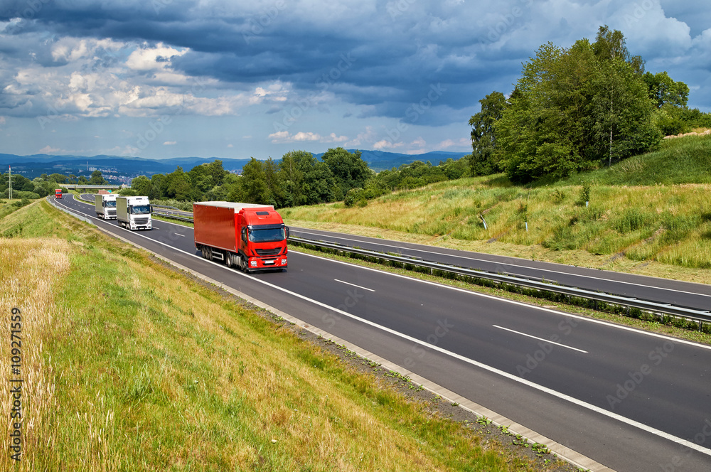 Landscape with an asphalt road and a row arriving red and white trucks. Forested mountains and city in the background in the distance. Sunny summer day with stormy clouds in the sky.