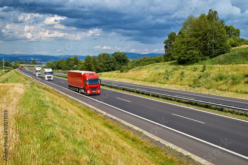 Obraz na płótnie Landscape with an asphalt road and a row arriving red and white trucks. Forested mountains and city in the background in the distance. Sunny summer day with stormy clouds in the sky. w salonie