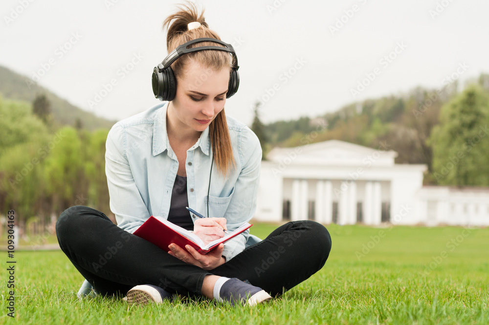 Fototapeta premium Young beautiful female sitting and writing outside with earphone