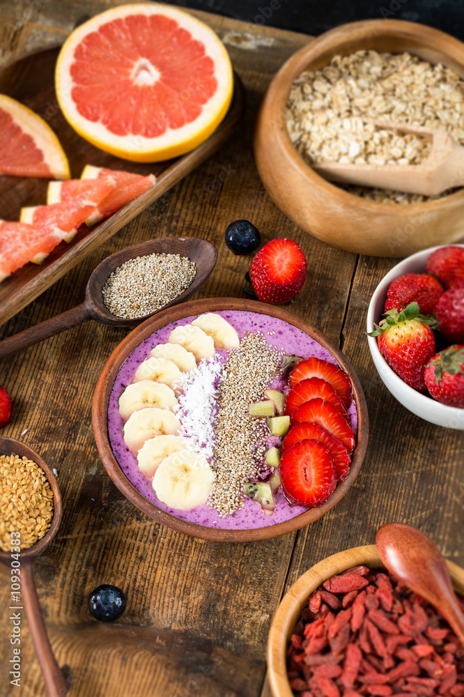 

Acai berry smoothie bowl topped with chia seeds, coconut flakes, banana, strawberries and kiwi with variety of superfoods on rustic wooden table. Healthy breakfast. Selective focus