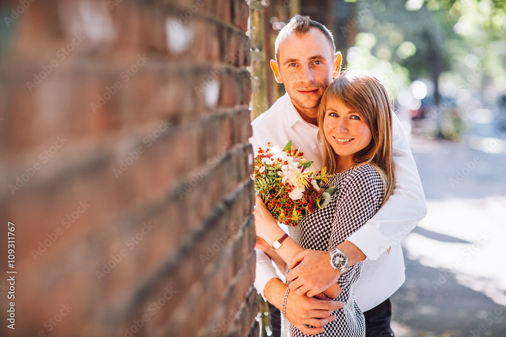 Fototapeta premium Close up fashion romantic portrait of young pretty couple kissing and hugs on the street at evening bright sunlight. Blonde gril with flowers boquet