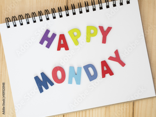 Word spelling "Happy Monday" on notebook page with wood background (Business concept)