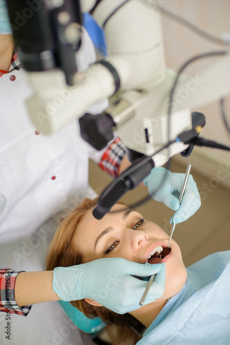 Portrait of young pretty female petient lying on dentist chair with open mouth. Dentist doctor treating root canals using microscope at dentistry office.