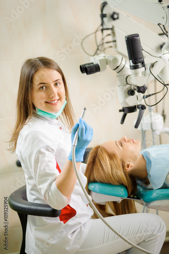 Portrait of beautiful female dentist holding stomatology tool  using microscope  smiling and looking at the camera at the dentist office. Dental care and treatment. Successful treatment