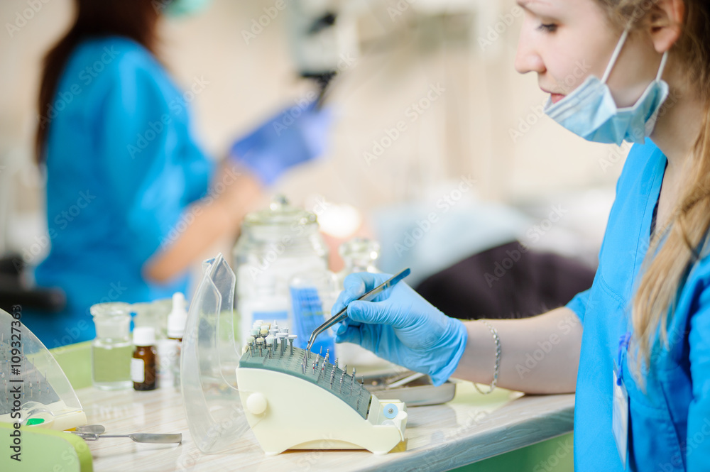 Close-up portrait of young female dentist assistant holding dental drill on foreground. Doctor working with microscope in the background. Dental equipment. Teeth care and tooth health Close-up portrait of young female dentist assistant holding dental drill on foreground. Doctor working with microscope in the background. Dental equipment. Teeth care and tooth health