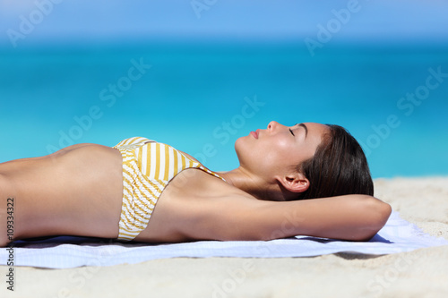 Summer beach vacation woman relaxing sunbathing and suntanning. Sexy young ethnic adult in yellow stripes bikini sleeping lying down on white sand dreaming on tropical travel tourist destination.