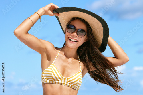 Summer beach vacation woman enjoying the sun in bikini and sunglasses having fun happy with hair in wind holding on hat. Sexy Asian young adult on towel for tropical travel holidays concept.