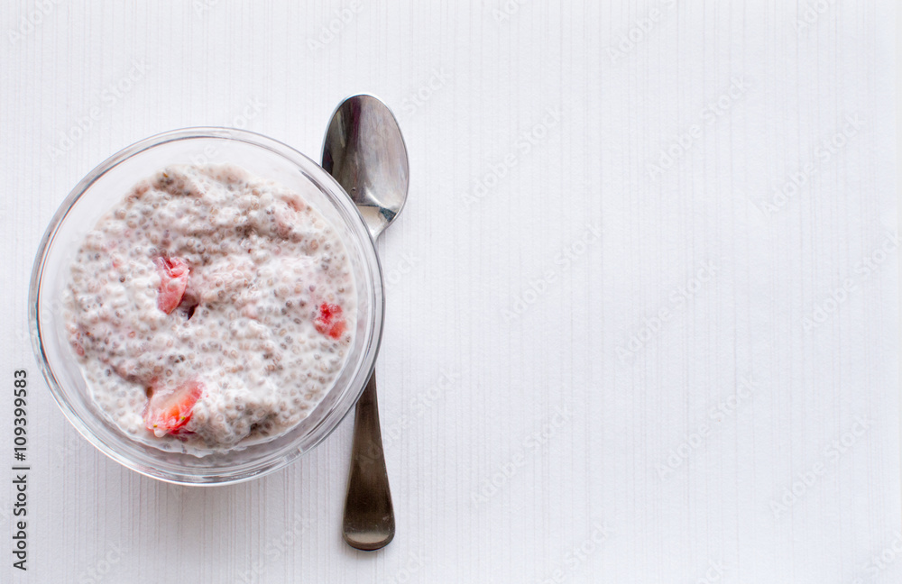 High angle view of chia seed and strawberry pudding in glass dish with spoon on white textured background