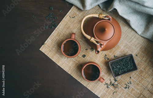 Chinese Tea Ceremony. Brown ceramic teapot and brown tea cup. Green tea on bamboo mat on aged wooden table. Top view.