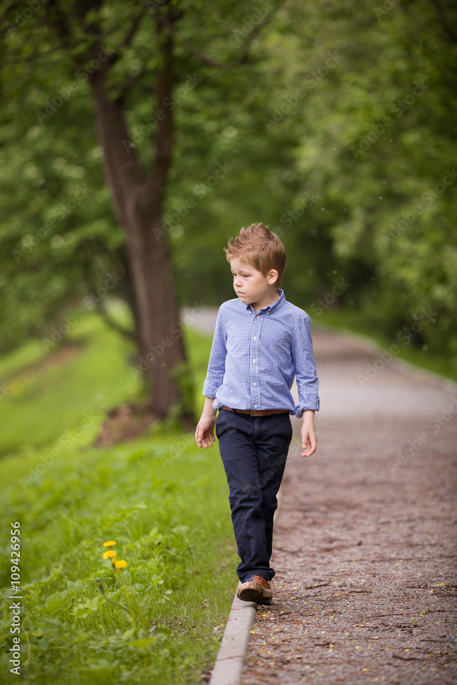 Cute little kid boy walking on the raod in the summer park. Summer forest background. School boy outdoors. Cute little kid boy walking on the raod in the summer park. Summer forest background. School boy outdoors.