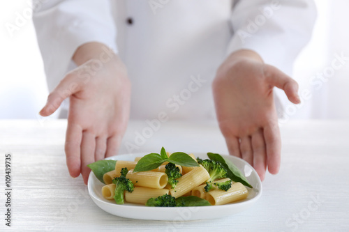 Female chef holding plate of boiled rigatoni pasta with broccoli and basil at the table