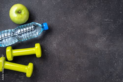 Fitness concept with dumbbells, water and green apple on dark background
