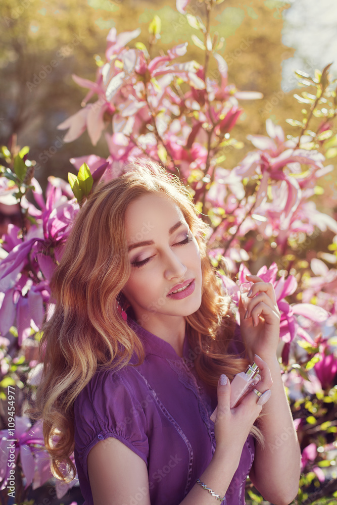 Outdoor portrait of a young beautiful  lady using perfume near magnolia tree with flowers. Model closed her eyes. Girl wearing stylish clothes. Female spring fashion
