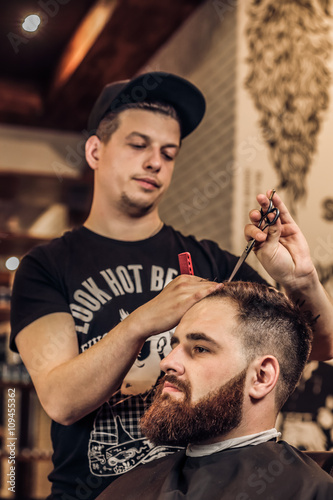 Closeup side view of young bearded man getting beard haircut by hairdresser at barbershop. Getting perfect shape while sitting in chair at barbershop. Grooming of real man.