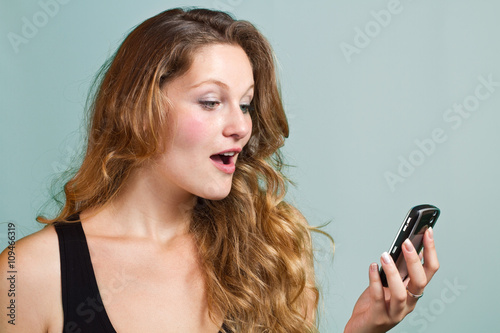 The young girl is holding a cell phone and looking at the screen with a surprised expression