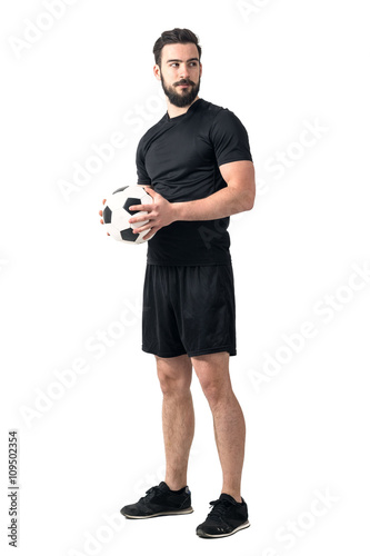 Strong confident soccer player holding ball with both hands looking away. Full body length portrait isolated over white background.