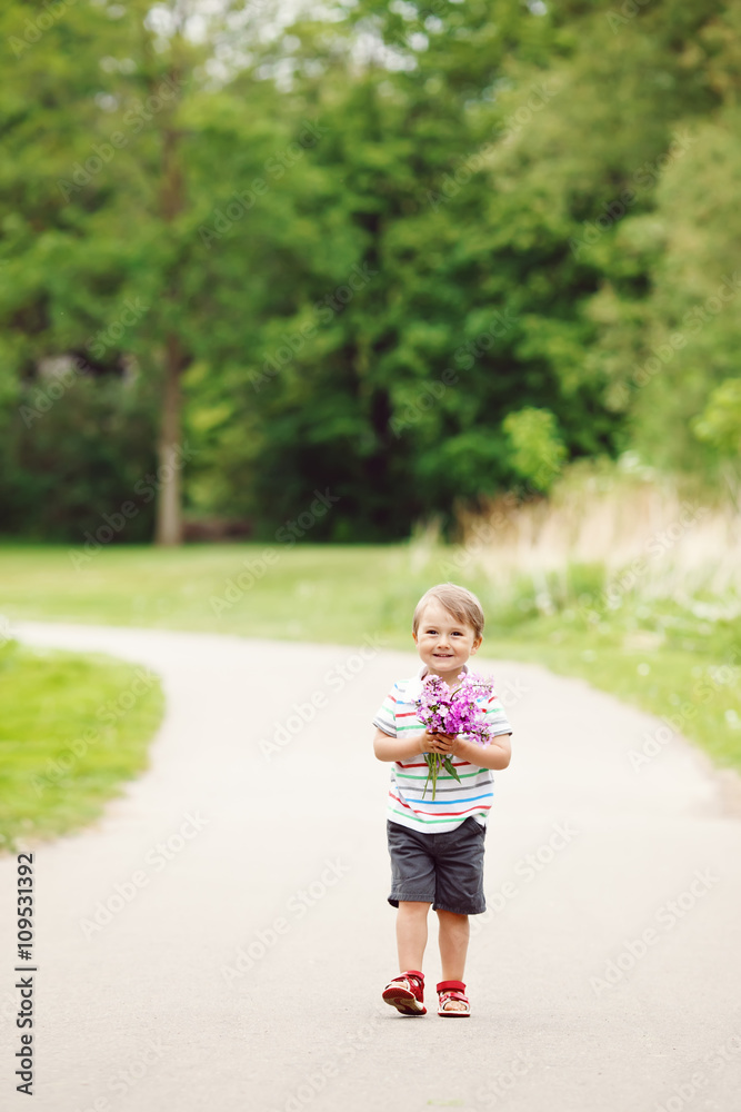 Portrait of a cute adorable funny little smiling boy toddler walking in park with lilac purple pink flowers in hands on bright summer day, mothers day holiday concept Portrait of a cute adorable funny little smiling boy toddler walking in park with lilac purple pink flowers in hands on bright summer day, mothers day holiday concept
