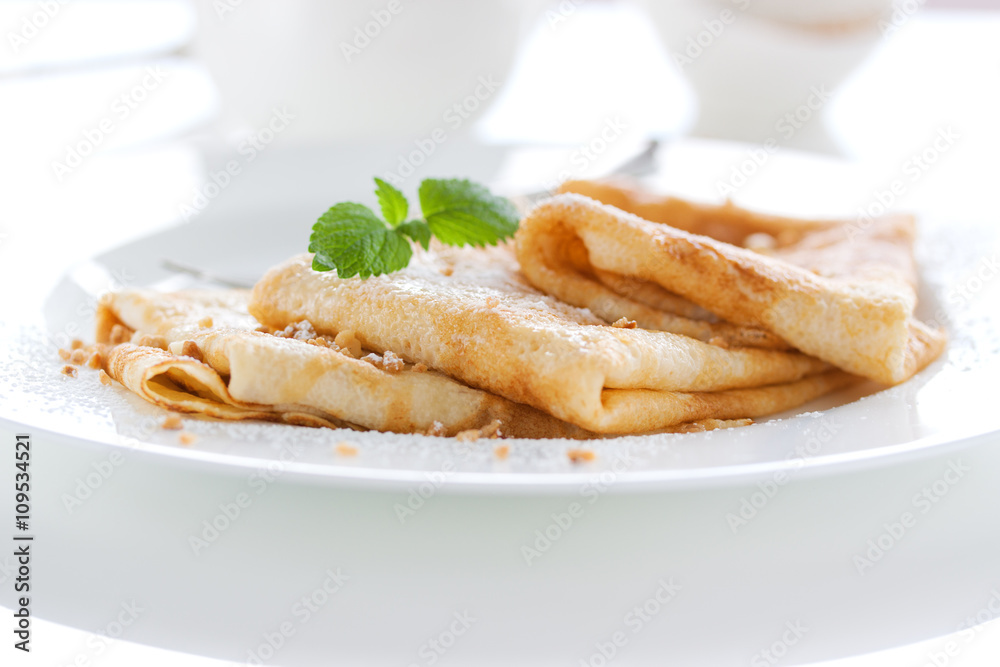 French crepes or pancakes with sugar powder, nuts and fresh mint on a white plate on a white background for breakfast, closeup