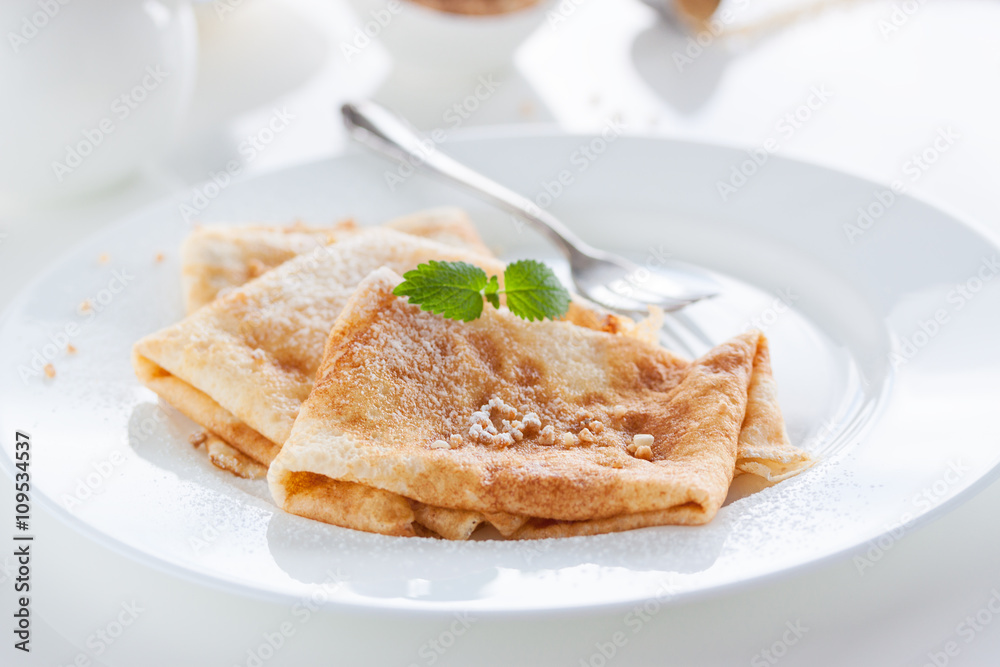 French crepes or pancakes with sugar powder, nuts and fresh mint on a white plate on a white background for breakfast, closeup