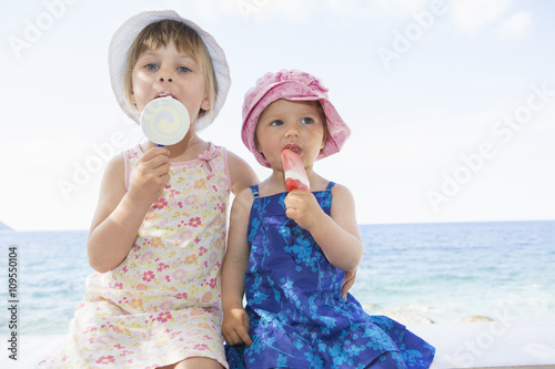 Female toddler and sister wearing sunhats eating ice lollies on beach photo