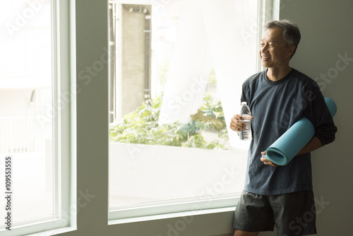 Mature man holding rolled up yoga mat and bottle of water looking out of window