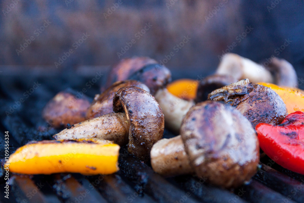 Seasoned mushrooms and peppers are being grilled on iron grates over charcoal Seasoned mushrooms and peppers are being grilled on iron grates over charcoal