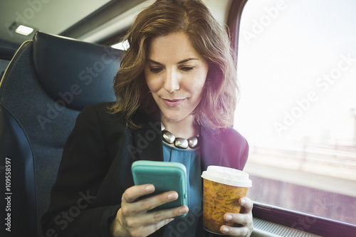 Mid adult woman on train, using smartphone, holding coffee cup photo
