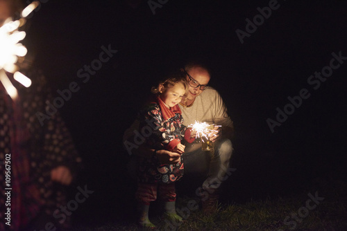 Father crouching next to daughter holding sparkler photo