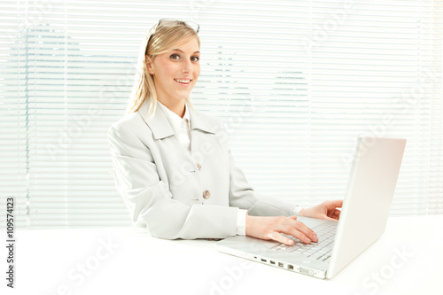 smiling blond businesswoman with laptop near venetian blind window