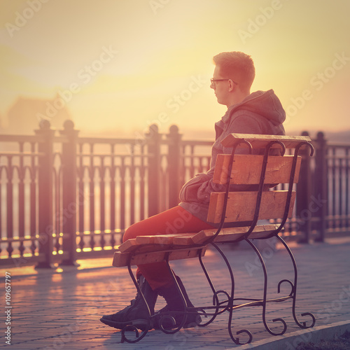 Photo of Lonely Man Sitting on a Bench during Sunset. Young Man Sitting on Wooden Bench. Sunset Male Outdoor Background