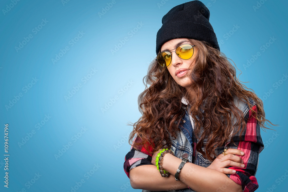 Cute hipster girl with long curly hair wearing red checkered shirt, denim vest, yellow glasses and black beanie hat posing at camera with crossed arms. Cute hipster girl with long curly hair wearing red checkered shirt, denim vest, yellow glasses and black beanie hat posing at camera with crossed arms.