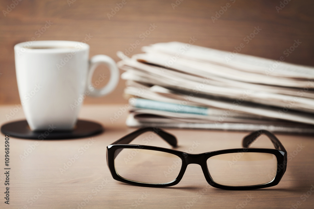Eyeglasses, coffee mug and stack of newspapers on wooden desk for themes of ophthalmology, poor vision and reading