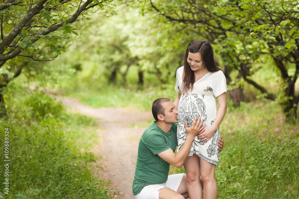 Fototapeta premium Happy couple waiting for baby. Future father kisses and hugs pregnant belly. Future mother is smiling and looking on man an her belly. New family concept. Paternity.