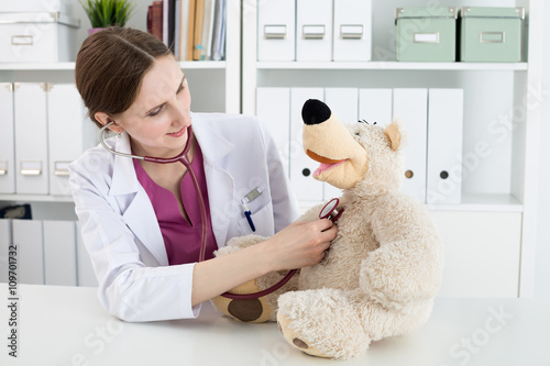 Beautiful smiling female doctor in white coat examine teddy bear