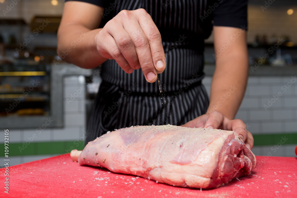 A fresh organic leg of lamb being seasoned with salt and pepper on a red chopping board.