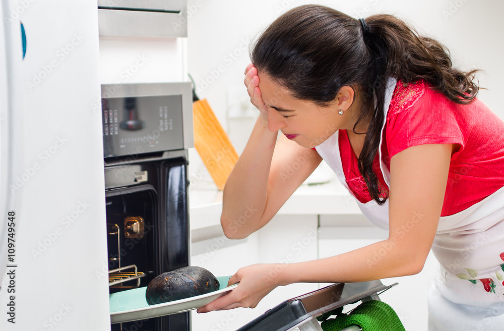 Young woman chef looking into oven with frustrated facial expression, holding black burnt bread on tray Young woman chef looking into oven with frustrated facial expression, holding black burnt bread on tray