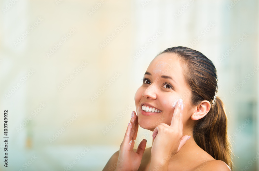 Pretty young healthy hispanic woman headshot with naked shoulders, smiling happily and applying cream to face using hands Pretty young healthy hispanic woman headshot with naked shoulders, smiling happily and applying cream to face using hands