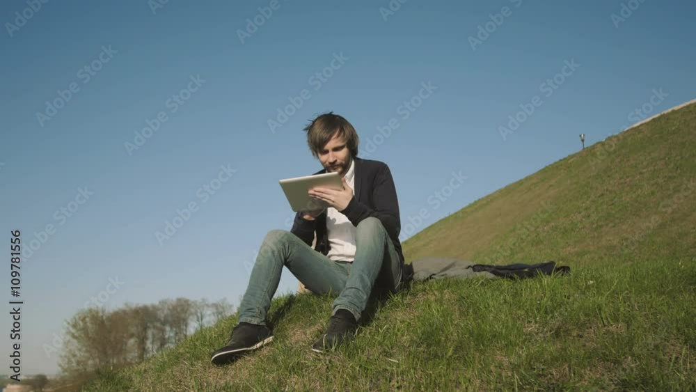 Handsome Hipster Using Tablet PC While Sitting On The Grass In The Park Handsome Hipster Using Tablet PC While Sitting On The Grass In The Park