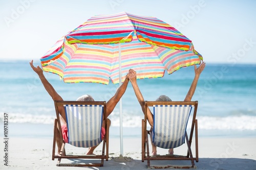 Photography Cute mature couple raising arms on deckchairs