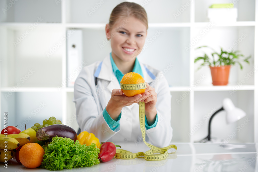 Smiling young female doctor measuring orange with tape sitting at the desk in the office. Concept of slimming, health and body care. Smiling young female doctor measuring orange with tape sitting at the desk in the office. Concept of slimming, health and body care.