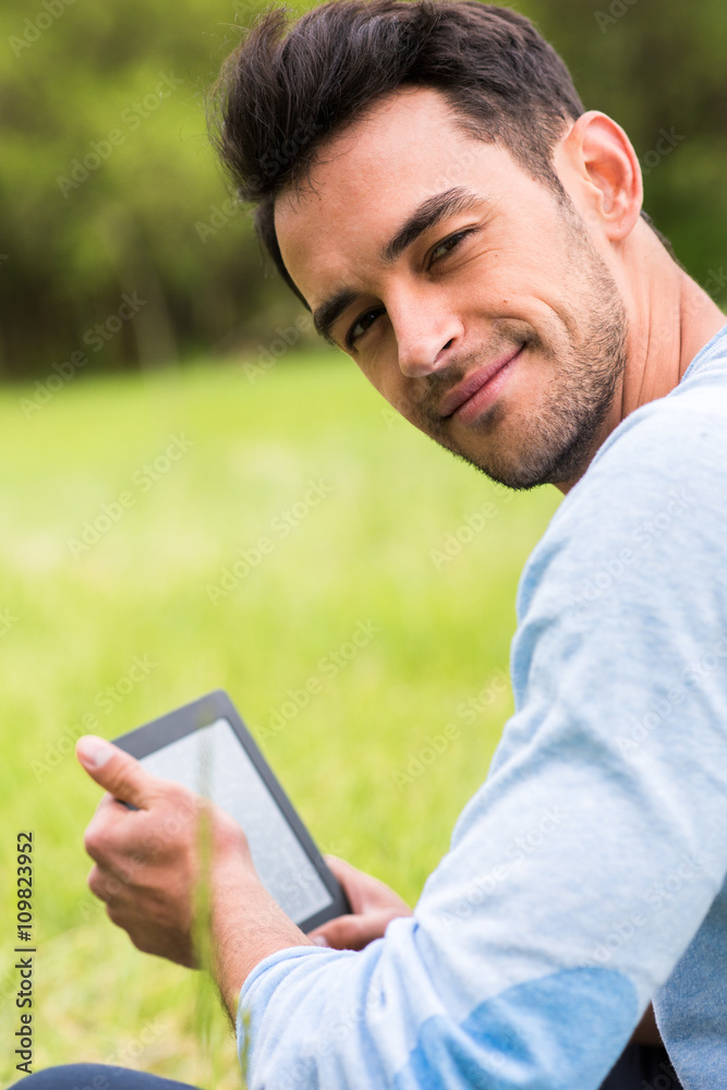Pretty smiling young business man with device look at the camera on nature green background Pretty smiling young business man with device look at the camera on nature green background