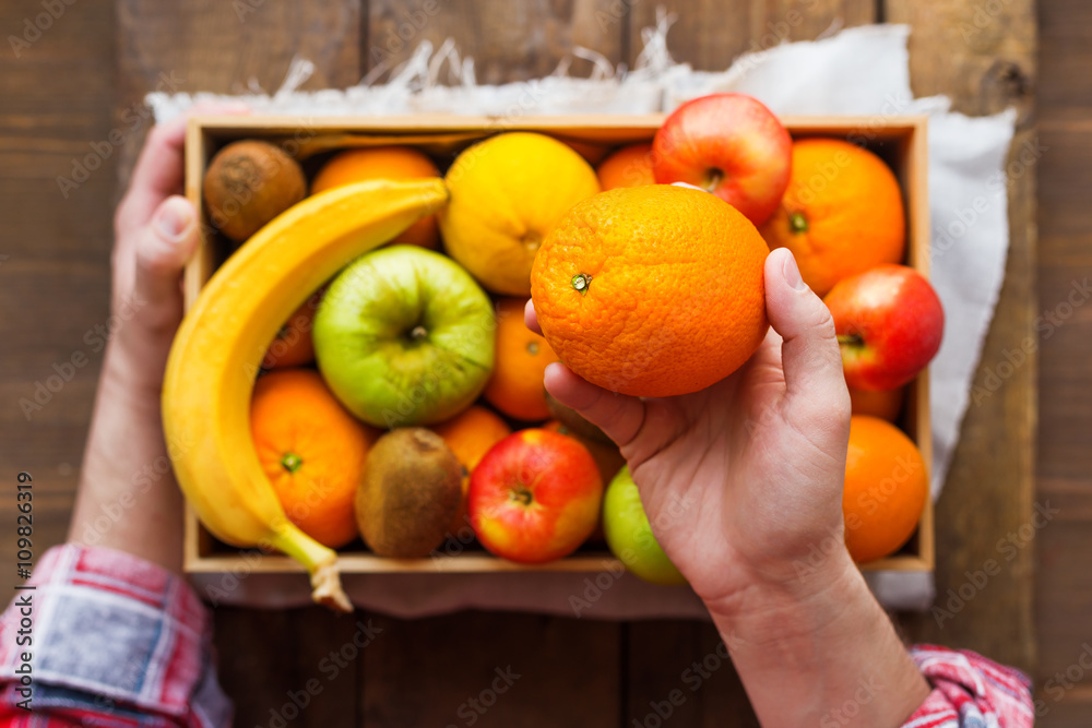 Man in tartan plaid shirt holds a box full of fresh fruits and an orange.