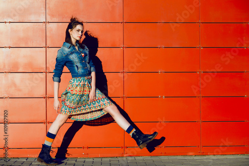 Young teenager girl posing, going, runs near urban red wall background in skirt and jeans jacket on the sunset. photo